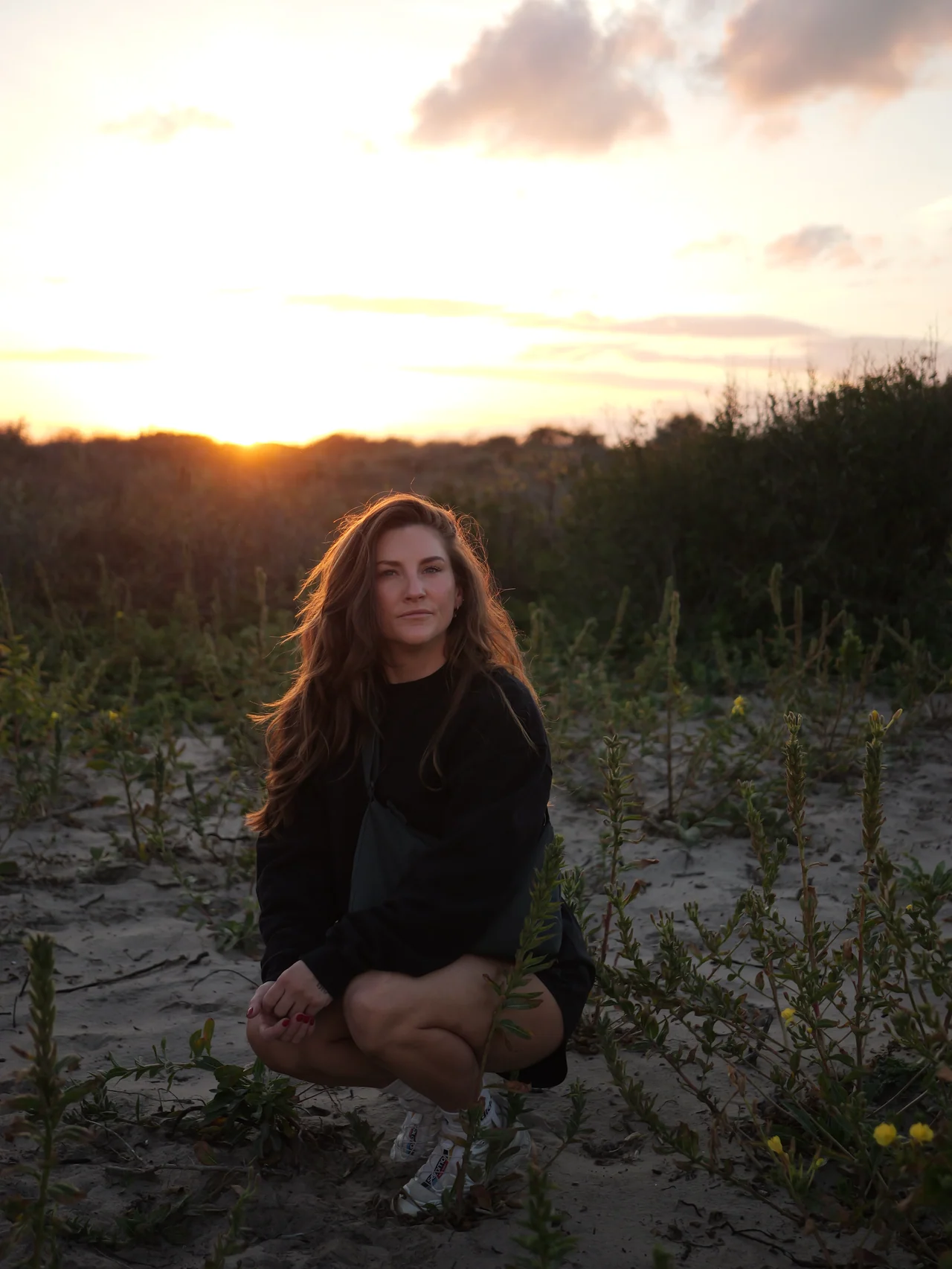 Een vrouw hurkt tussen de duinplanten bij zonsondergang
